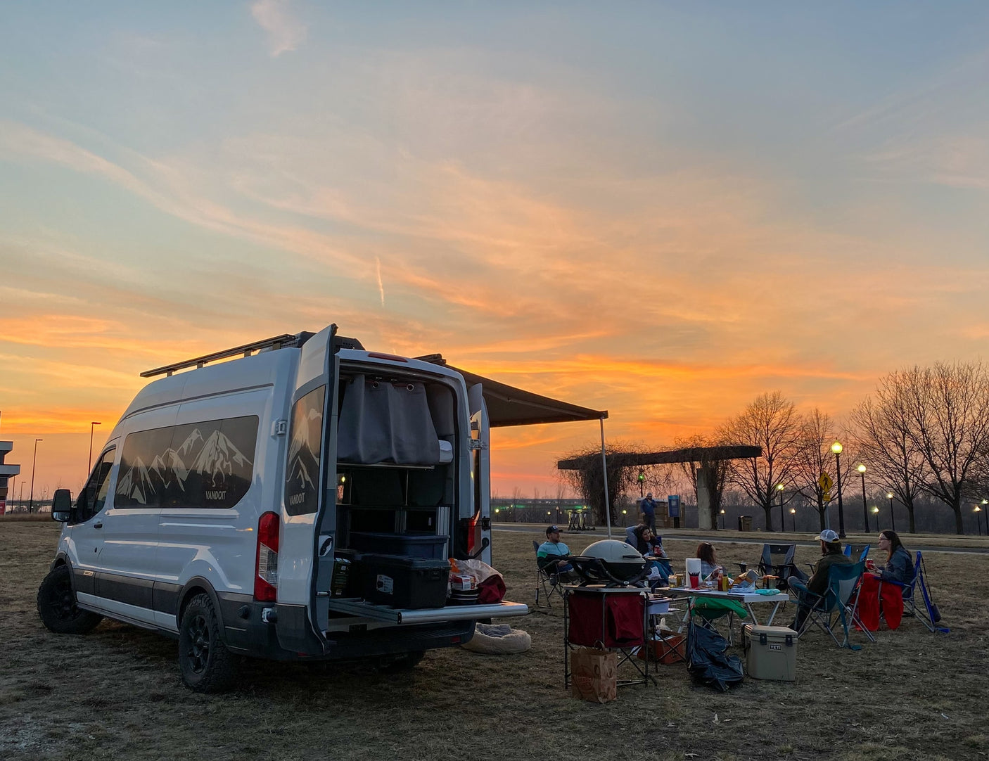Vibrant sunset view: Friends enjoy outdoor cooking by a Vandoit RV rental camper van.|Friends and dogs relax by an RV rental van with an extended awning in a park