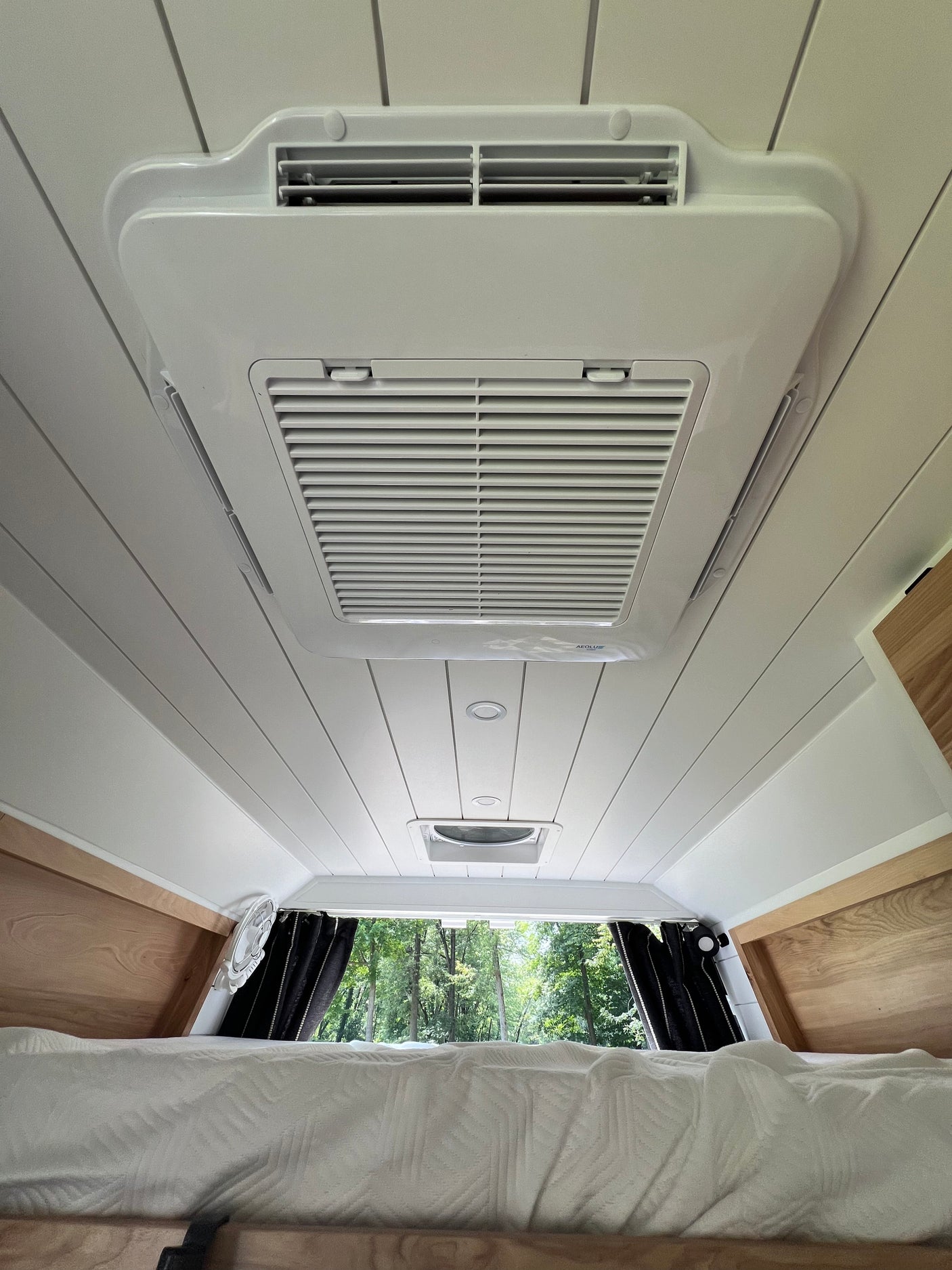 Inside a spacious RV rental, featuring its ceiling-mounted AC unit, vent, and comfortable bed with a forest view.
