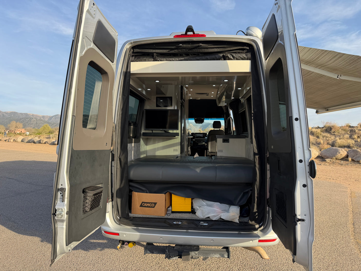Open rear doors of a silver RV rental, showing the interior with a bed, TV, and storage, ready for adventure.