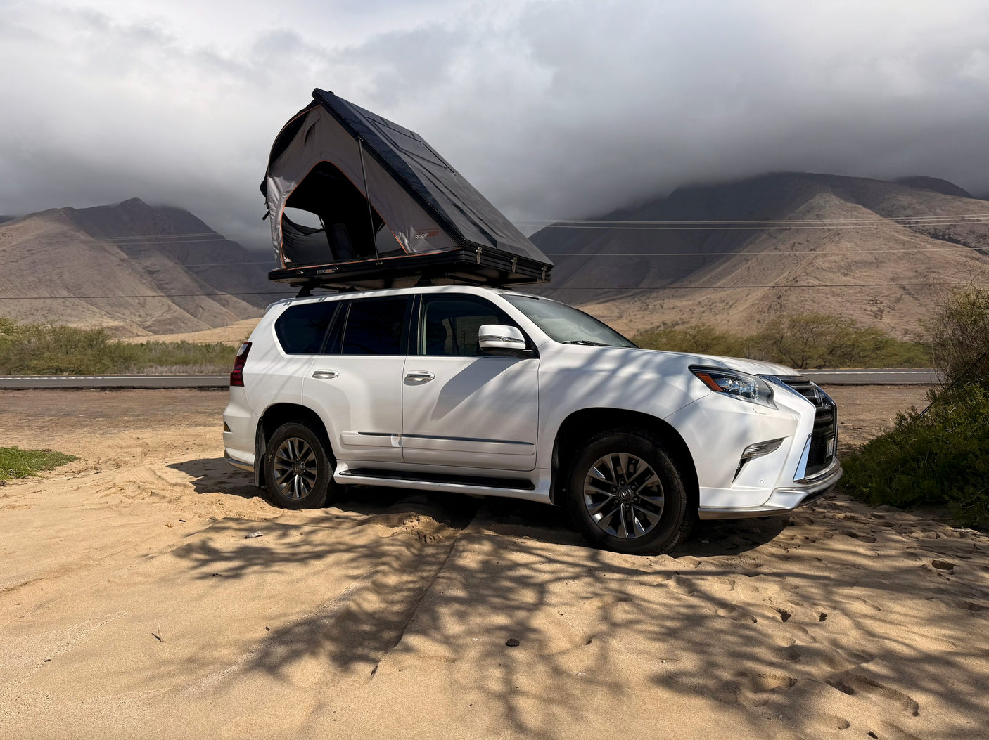 White Lexus GX SUV with hardshell rooftop tent, parked on sand with desert mountains & cloudy sky. Great RV rental option.