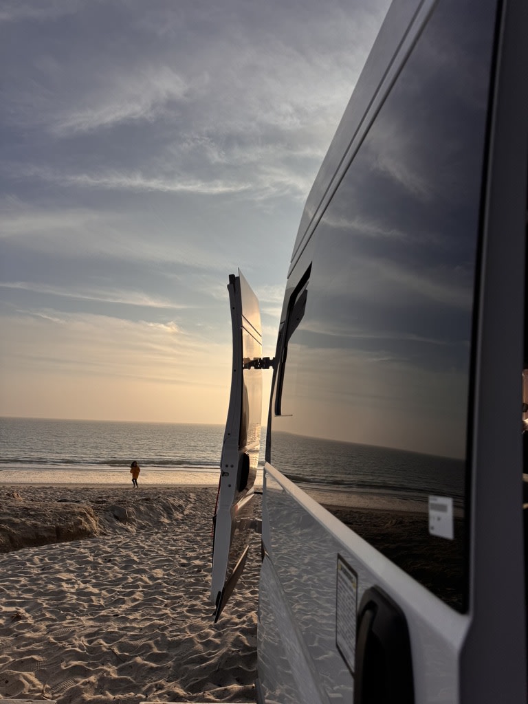 Scenic rv rental parked on a sandy beach. Open van door offers a view of the ocean, soft sunset, and a distant person.