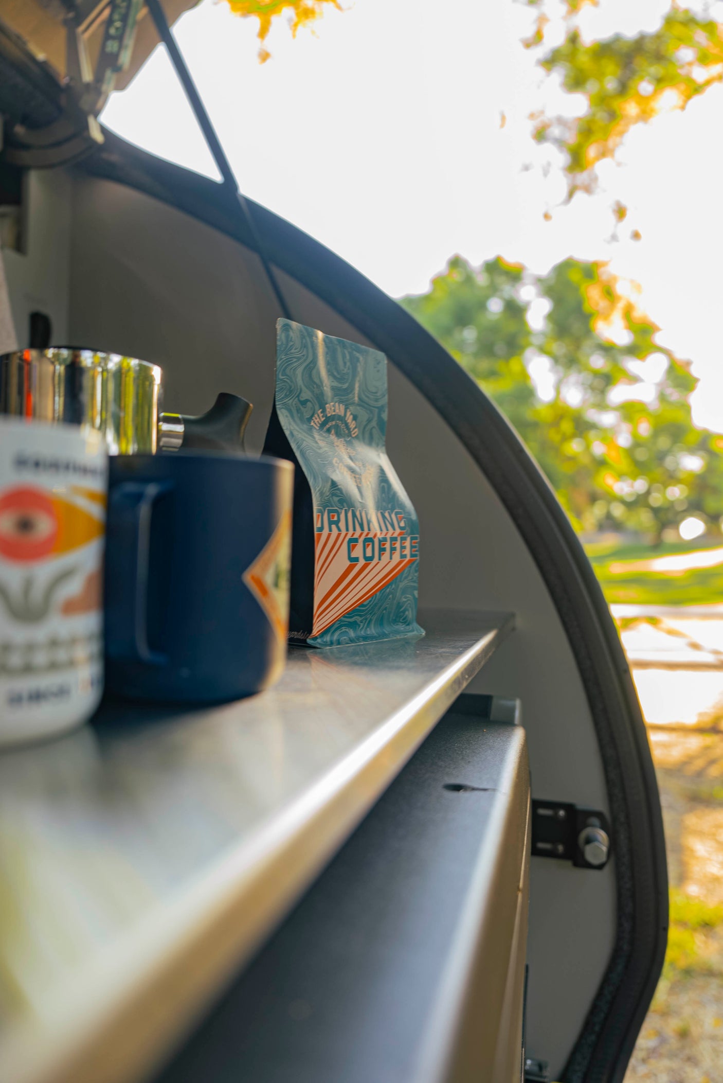 A close-up of coffee supplies inside an rv rental: a white mug, blue mug, metal pot, and a "DRINKING COFFEE" bag on a shelf. Outdoors view.
