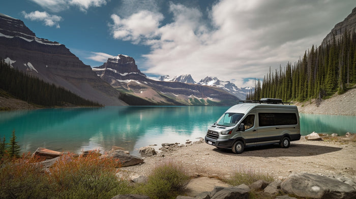 A light gray RV rental camper van is parked on a gravel shore beside a vivid turquoise lake, surrounded by snow-capped mountains and evergreen forests.