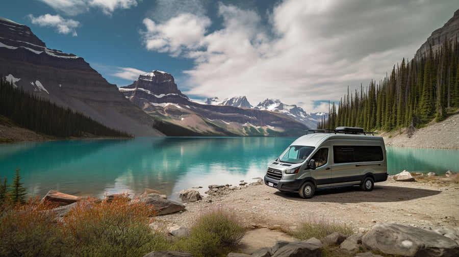A light gray RV rental camper van is parked on a gravel shore beside a vivid turquoise lake, surrounded by snow-capped mountains and evergreen forests.