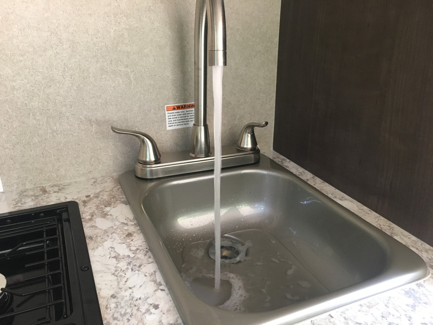 This RV rental kitchen features a stainless steel sink with water running from the faucet, and a black cooktop on the left.
