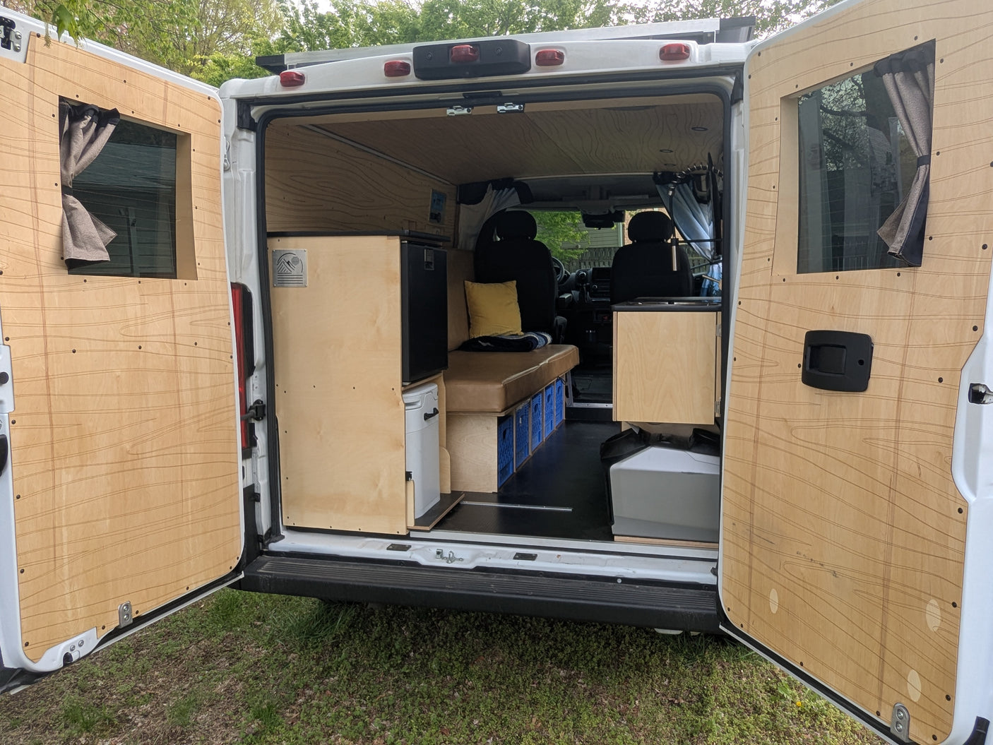 Open rear view of a custom-built RV rental interior. Features wood paneling, fridge, portable toilet, bench, and storage.