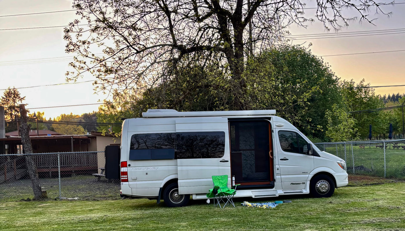 A white Pleasure-Way camper van, a perfect RV rental, parked on a grassy lawn at sunset with a chair and blanket for outdoor leisure.