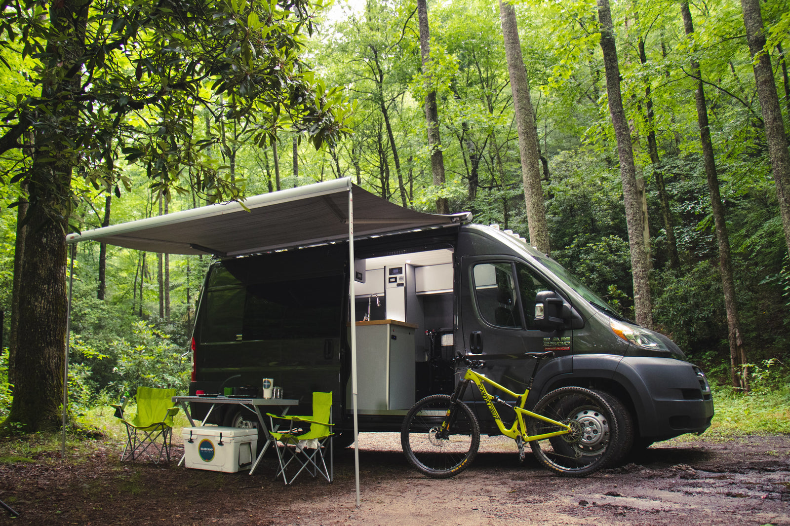 An RV rental camper van parked in a lush forest, featuring an extended awning, outdoor seating, and a mountain bike ready for adventure.