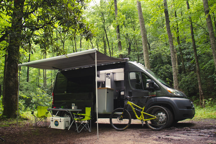 An RV rental camper van parked in a lush forest, featuring an extended awning, outdoor seating, and a mountain bike ready for adventure.