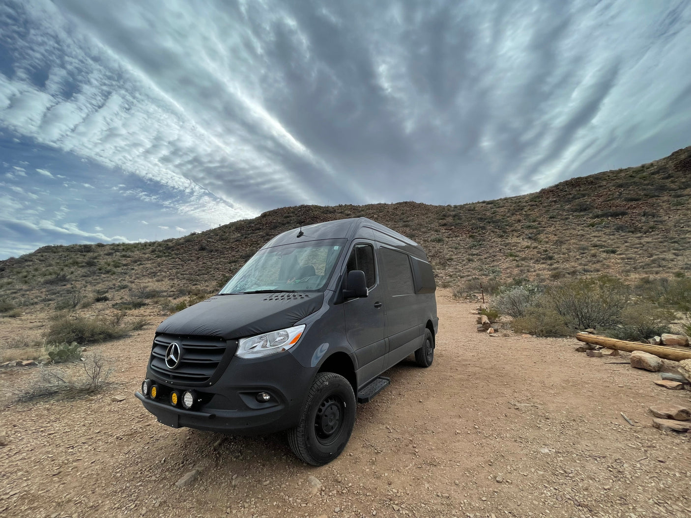 Experience desert adventures! A dark gray Sprinter camper van, perfect for RV rental, parked on a dusty trail with dramatic clouds.