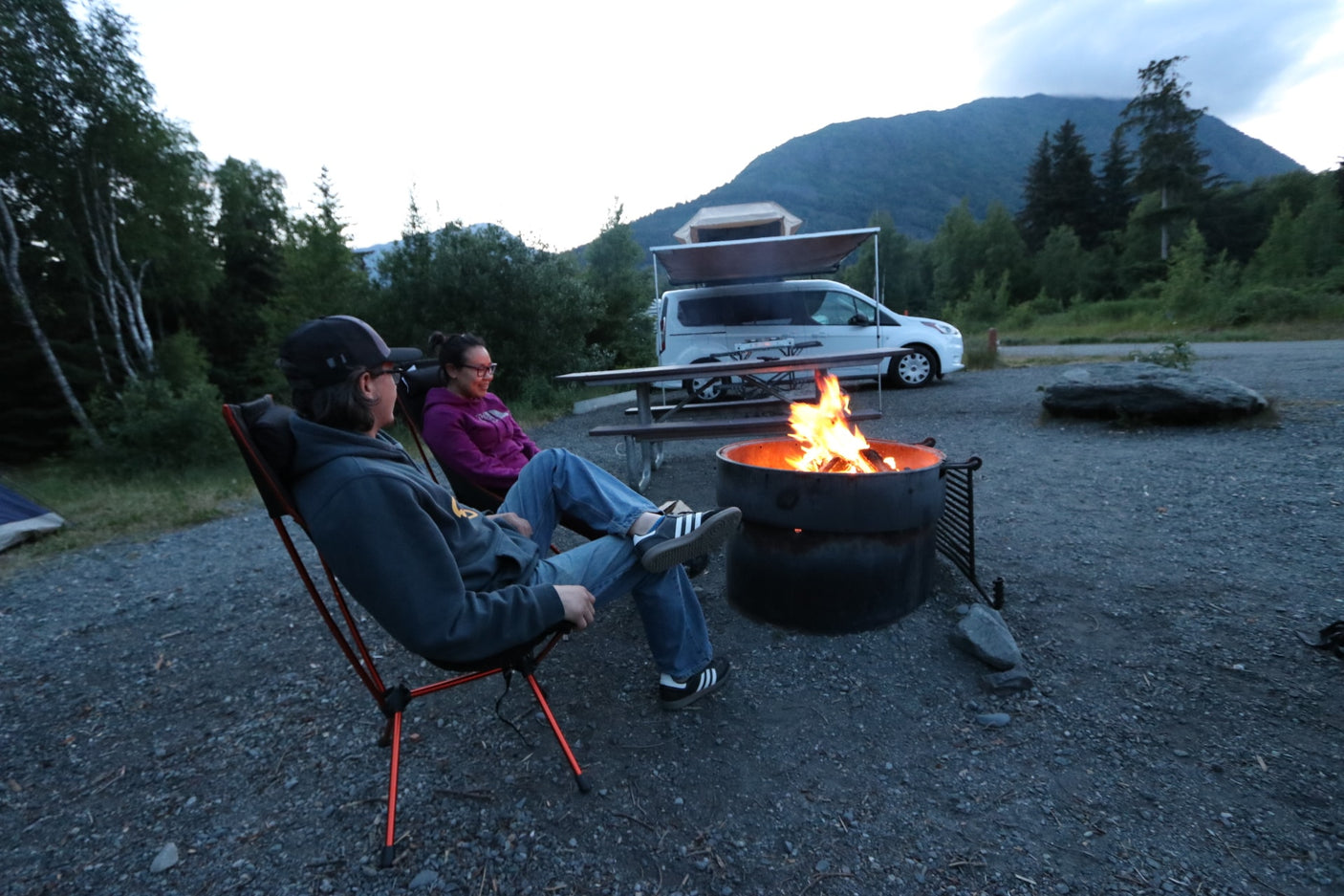 Two people relax by a campfire at dusk, enjoying a scenic campsite with an RV rental and mountains.