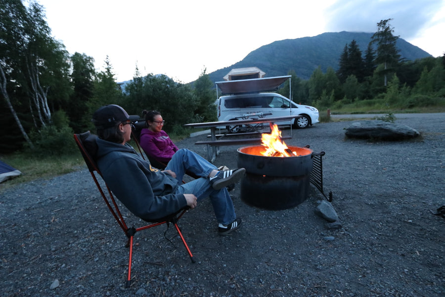 Two people relax by a campfire at dusk, enjoying a scenic campsite with an RV rental and mountains.