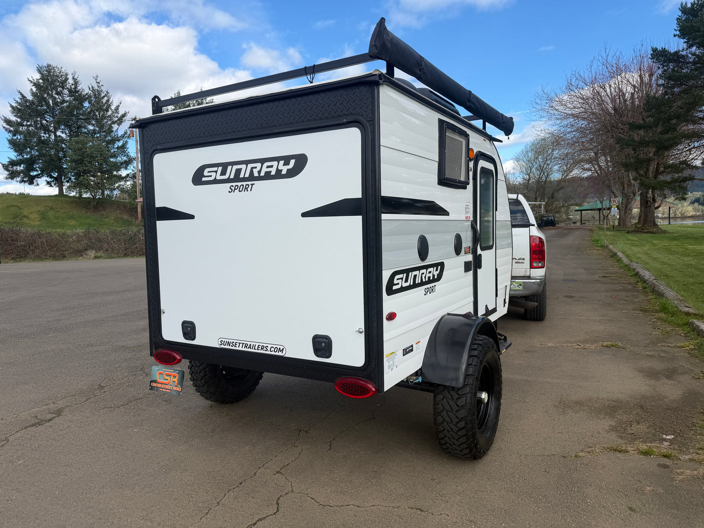 White SUNRAY SPORT teardrop RV rental with off-road tires, roof rack, and awning, hitched to a truck outdoors.