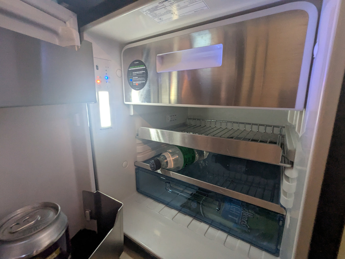 RV rental Dometic compact fridge interior, well-lit with a bottle, can, and shelving for your chilled refreshments.