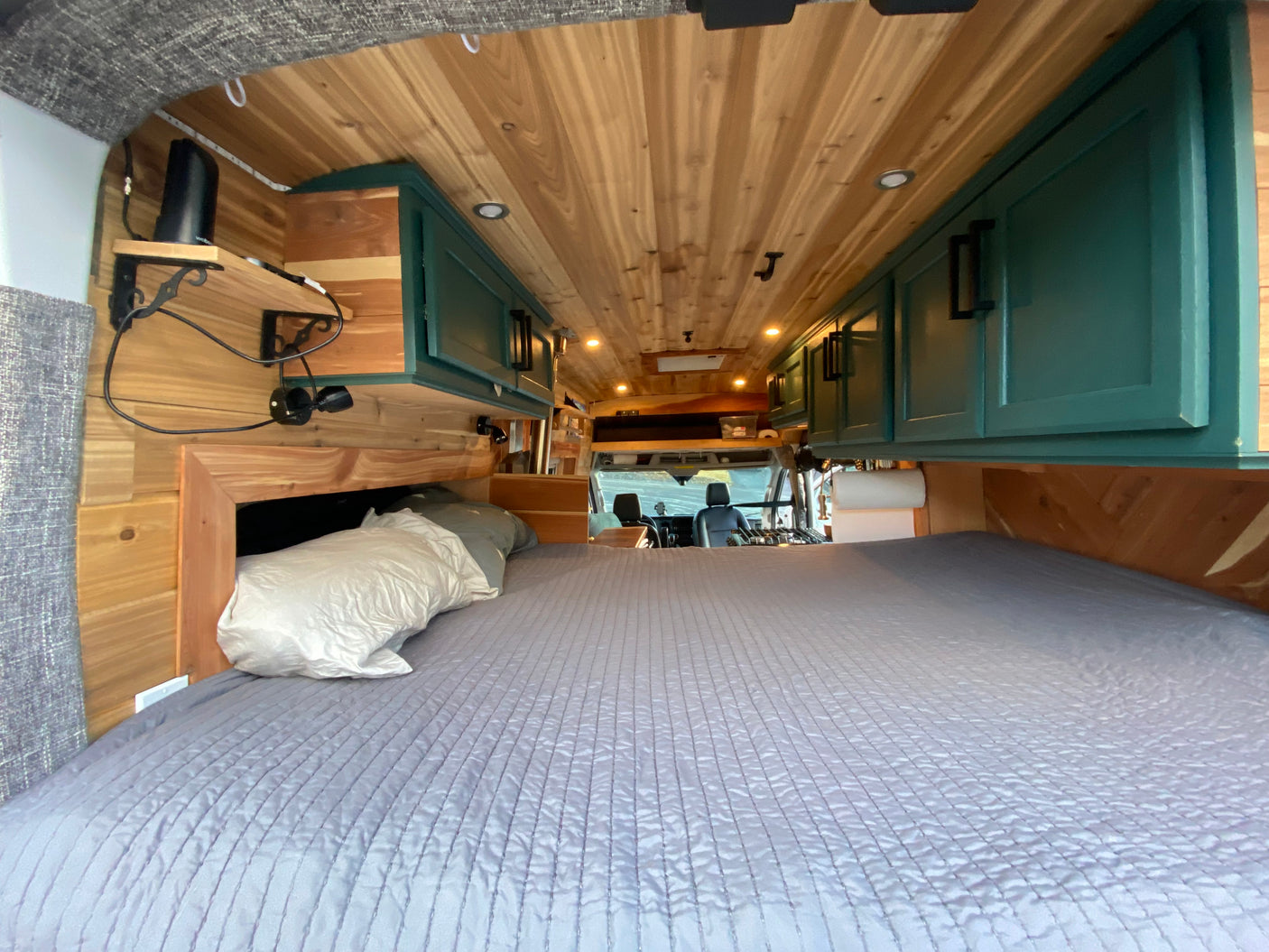 An RV rental's inviting sleeping area featuring a quilted bed, warm wood paneling, and teal overhead storage cabinets.
