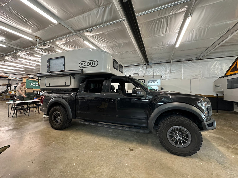 Black Ford Raptor pickup with a "Scout" truck camper, perfect for overland RV rental adventures. In a brightly lit workshop.