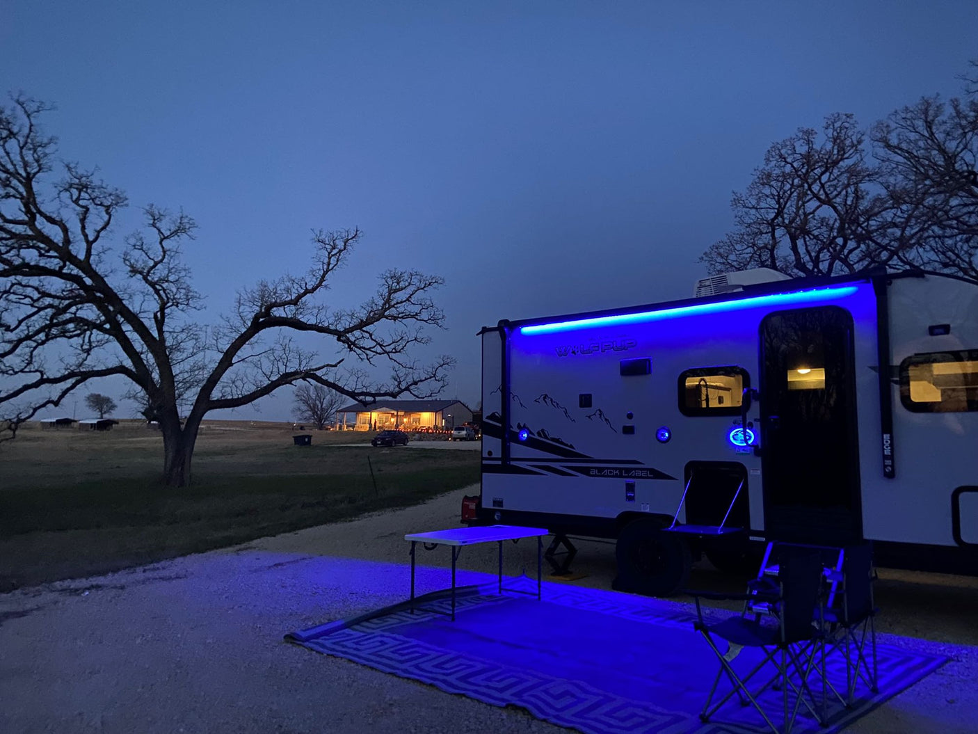Enjoying the evening at an RV rental campsite. Blue lights glow on the trailer, outdoor rug, table & chairs.