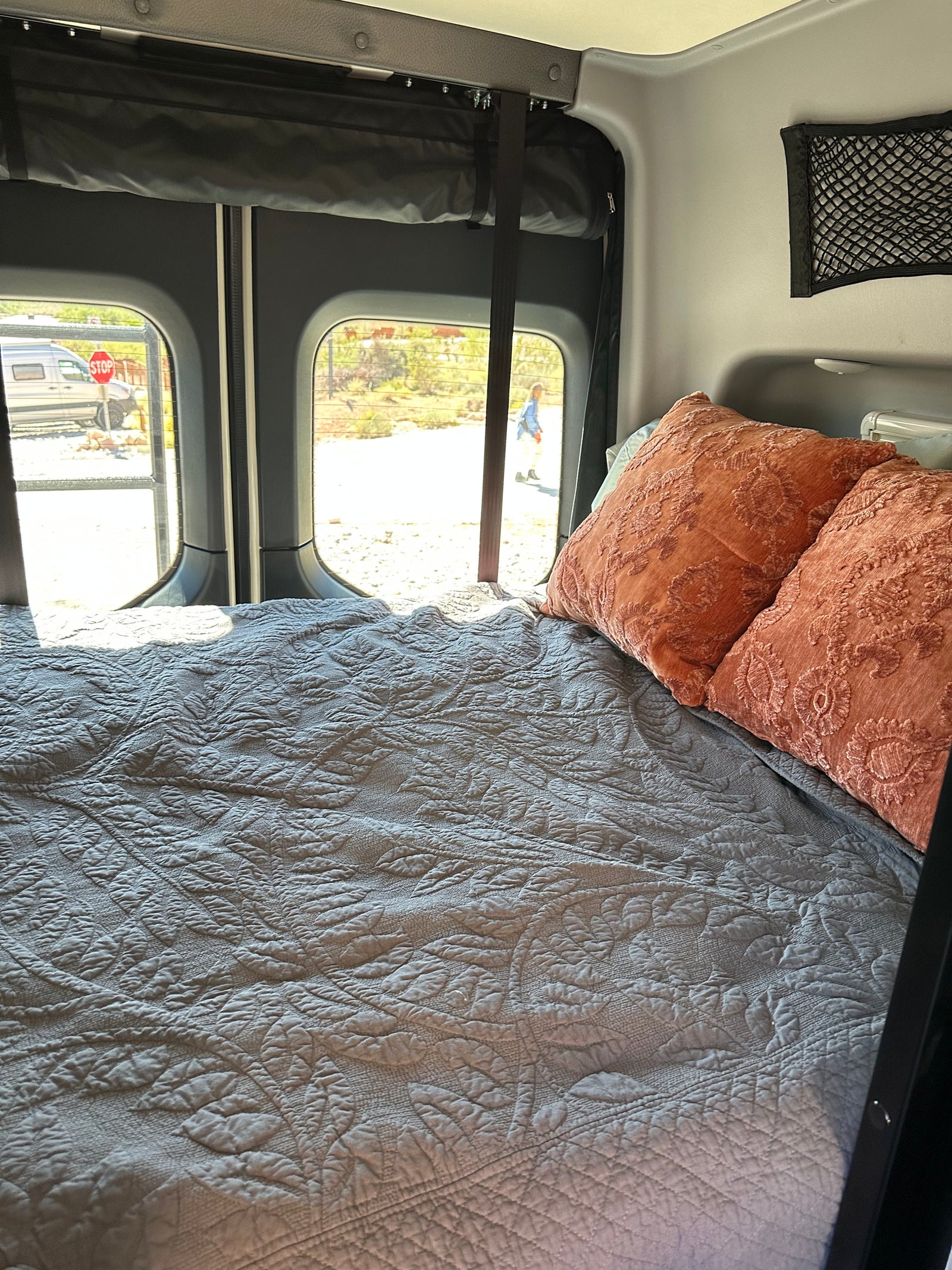 A comfortable bed with a gray quilted cover and orange pillows inside an rv rental, with sunny outdoor views.