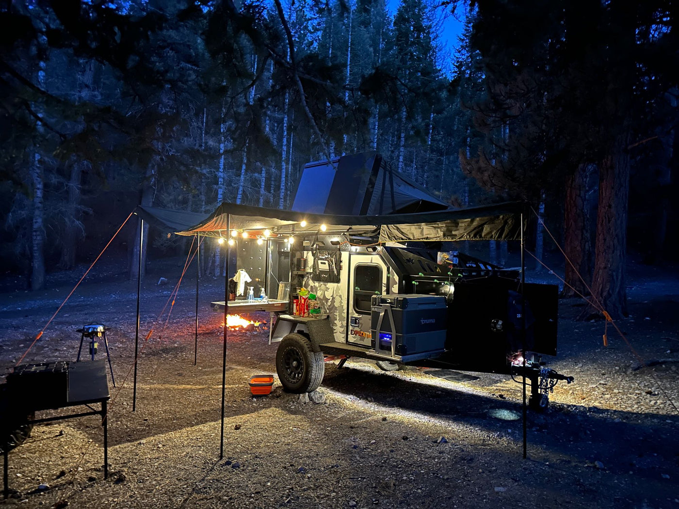 An off-road trailer RV rental lights up a cozy nighttime campsite in the forest, featuring a warm campfire and outdoor gear. — photo 18