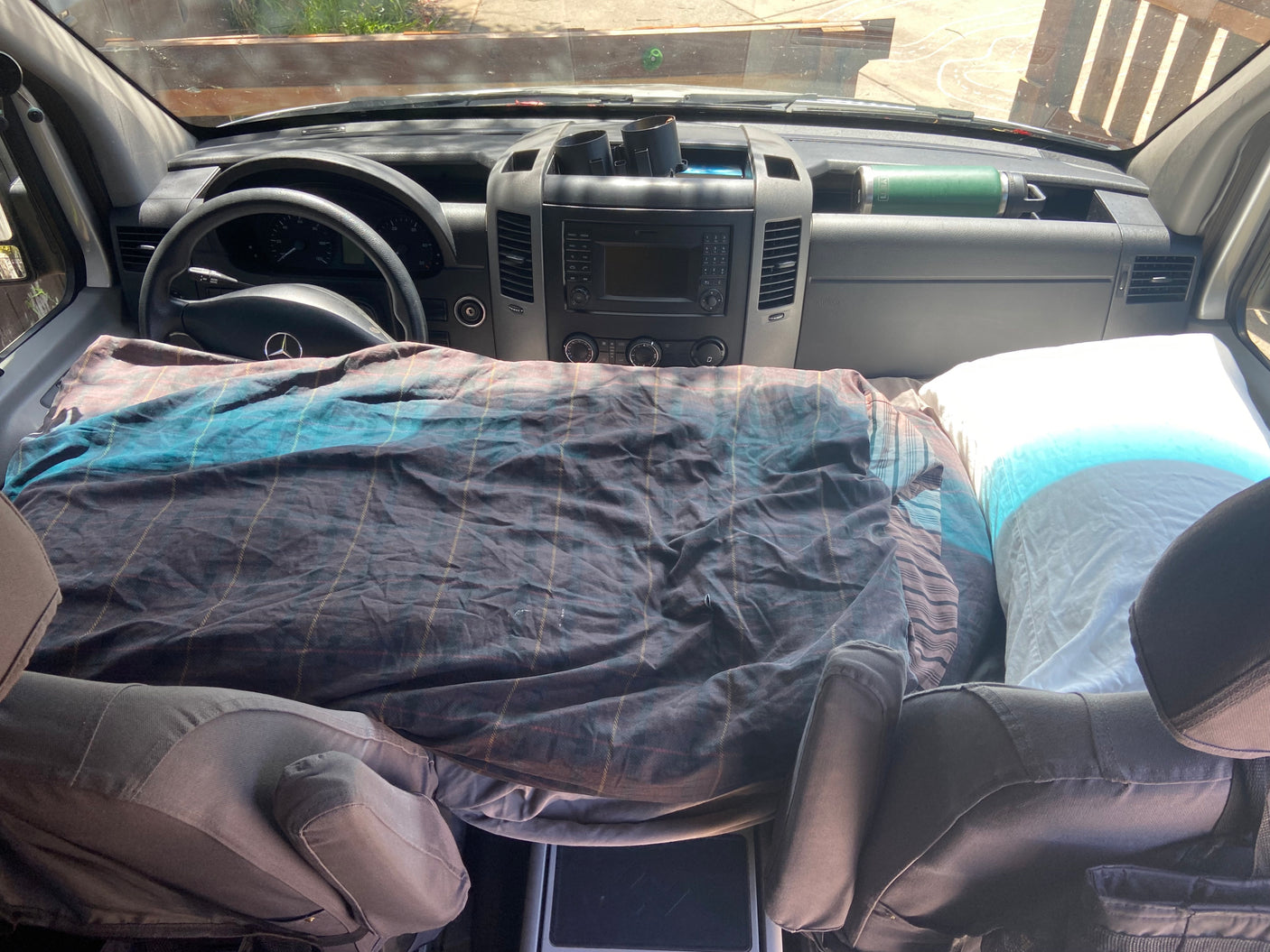 RV rental interior: a cozy bed with plaid bedding fills the front cabin, featuring a Mercedes dashboard and Yeti bottle.