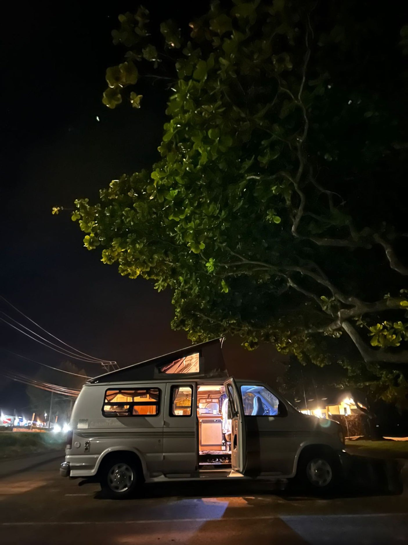 Night scene: a spacious pop-top RV rental with interior lights on and door open, parked under a large tree.