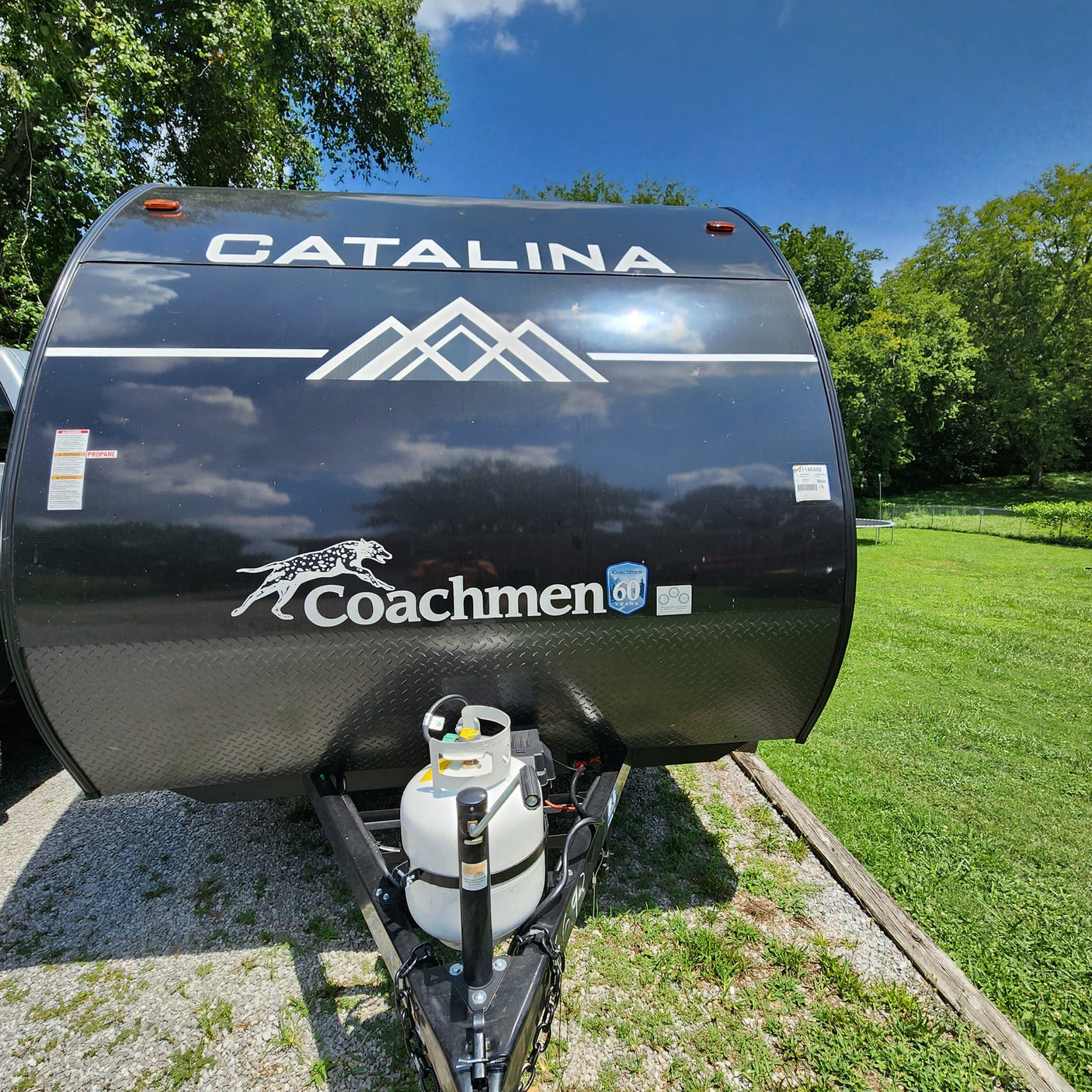 Front view of a black Catalina Coachmen RV rental travel trailer with propane tanks, parked on gravel in a sunny outdoor setting.