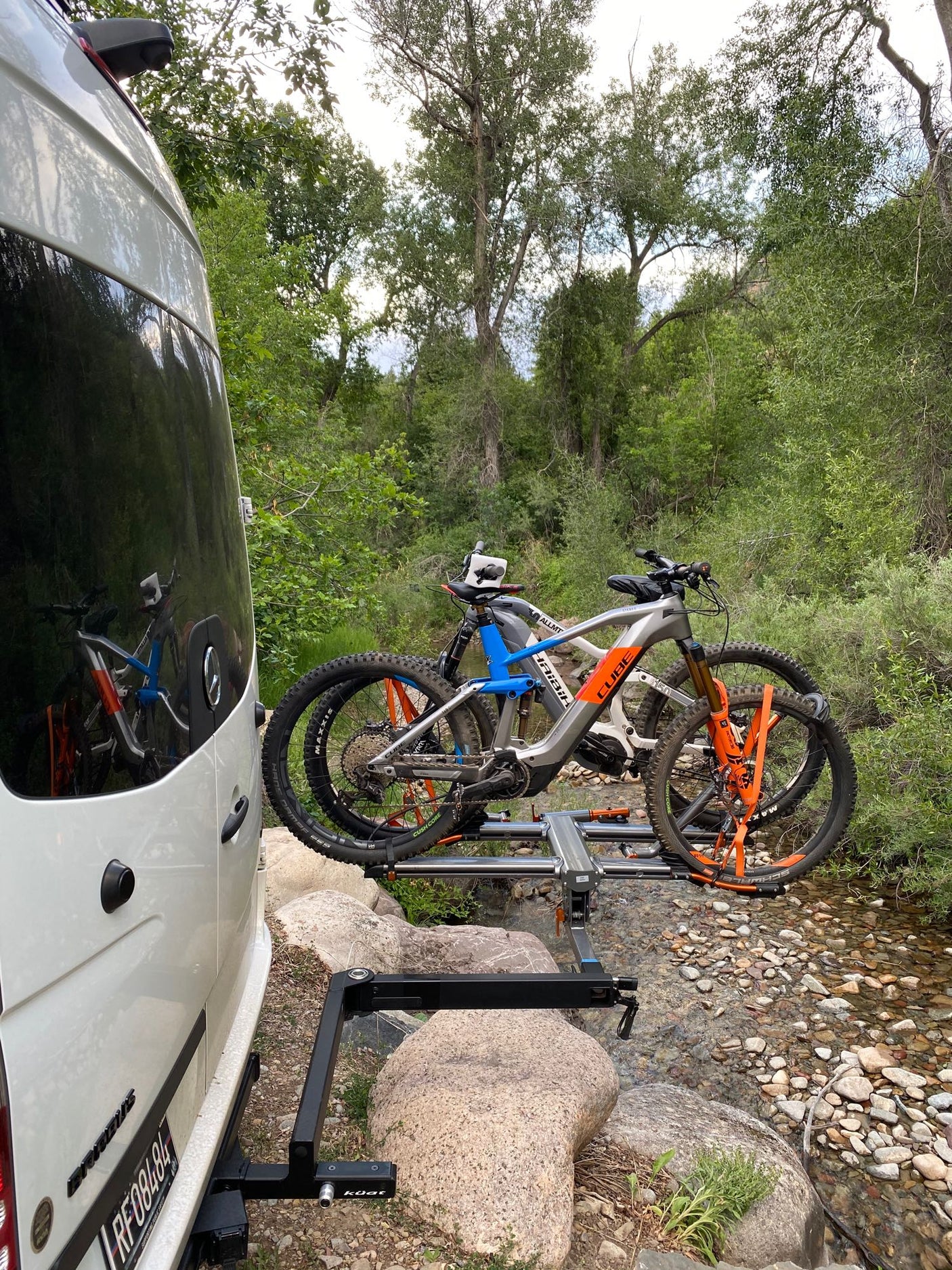 RV rental with two electric mountain bikes on a rack, parked beside a rocky stream, ready for outdoor adventure.