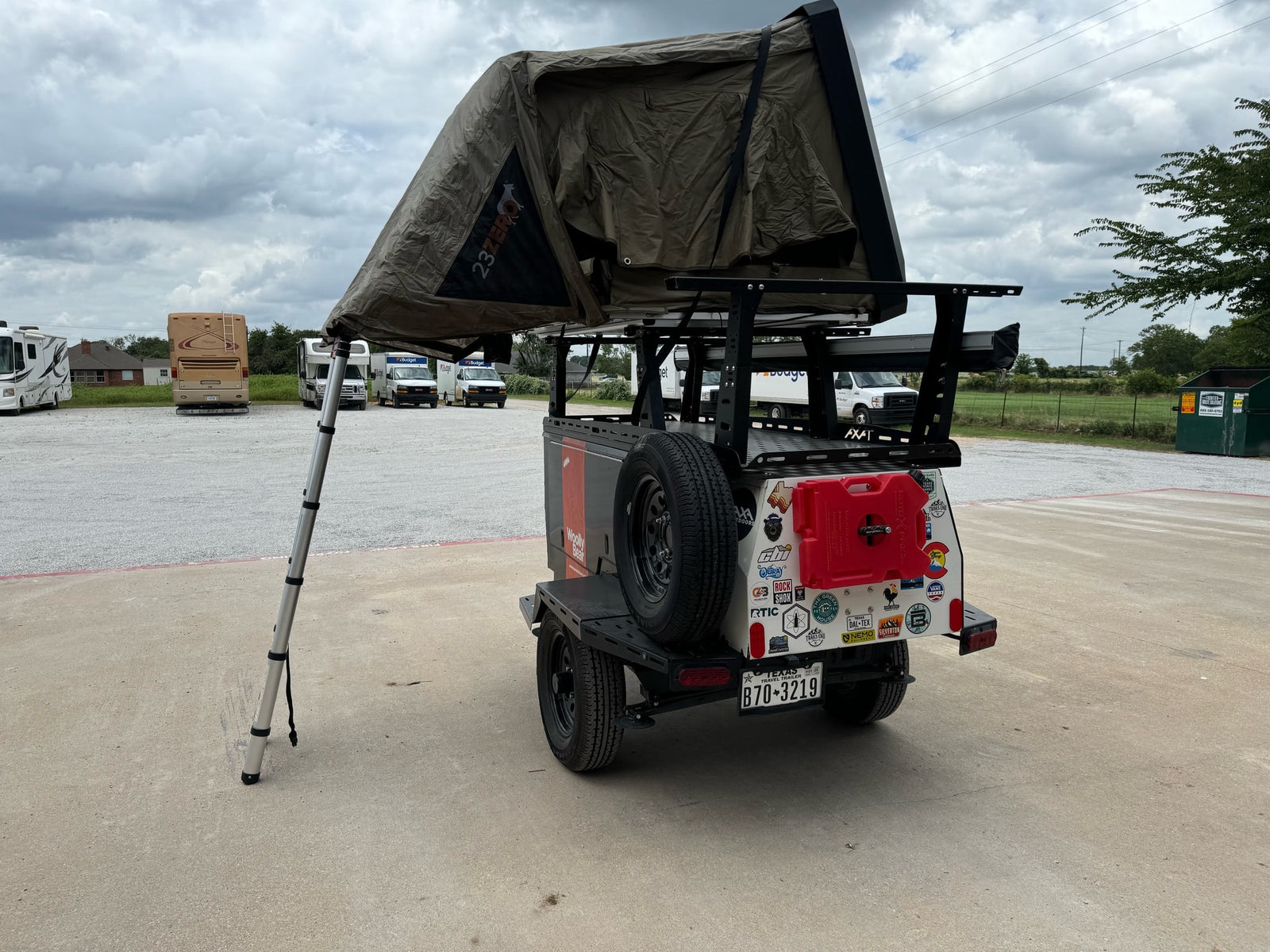 An equipped camping trailer with a 23ZERO rooftop tent displayed at an RV rental lot, surrounded by RVs and rental vans.