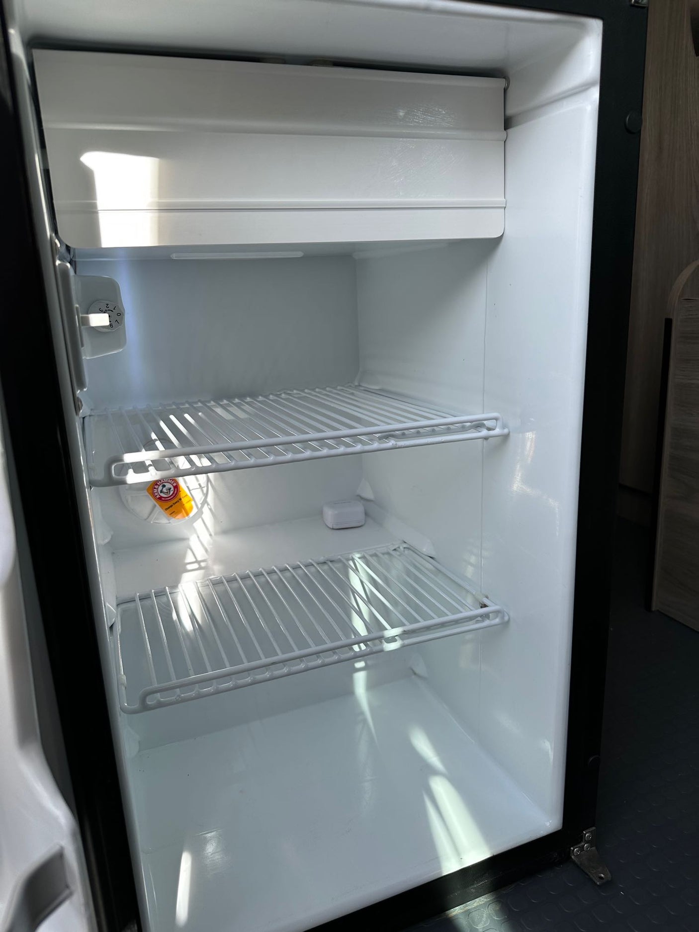 An open, clean, and empty RV rental mini-fridge, displaying wire shelves, a small freezer, and a thermostat dial.