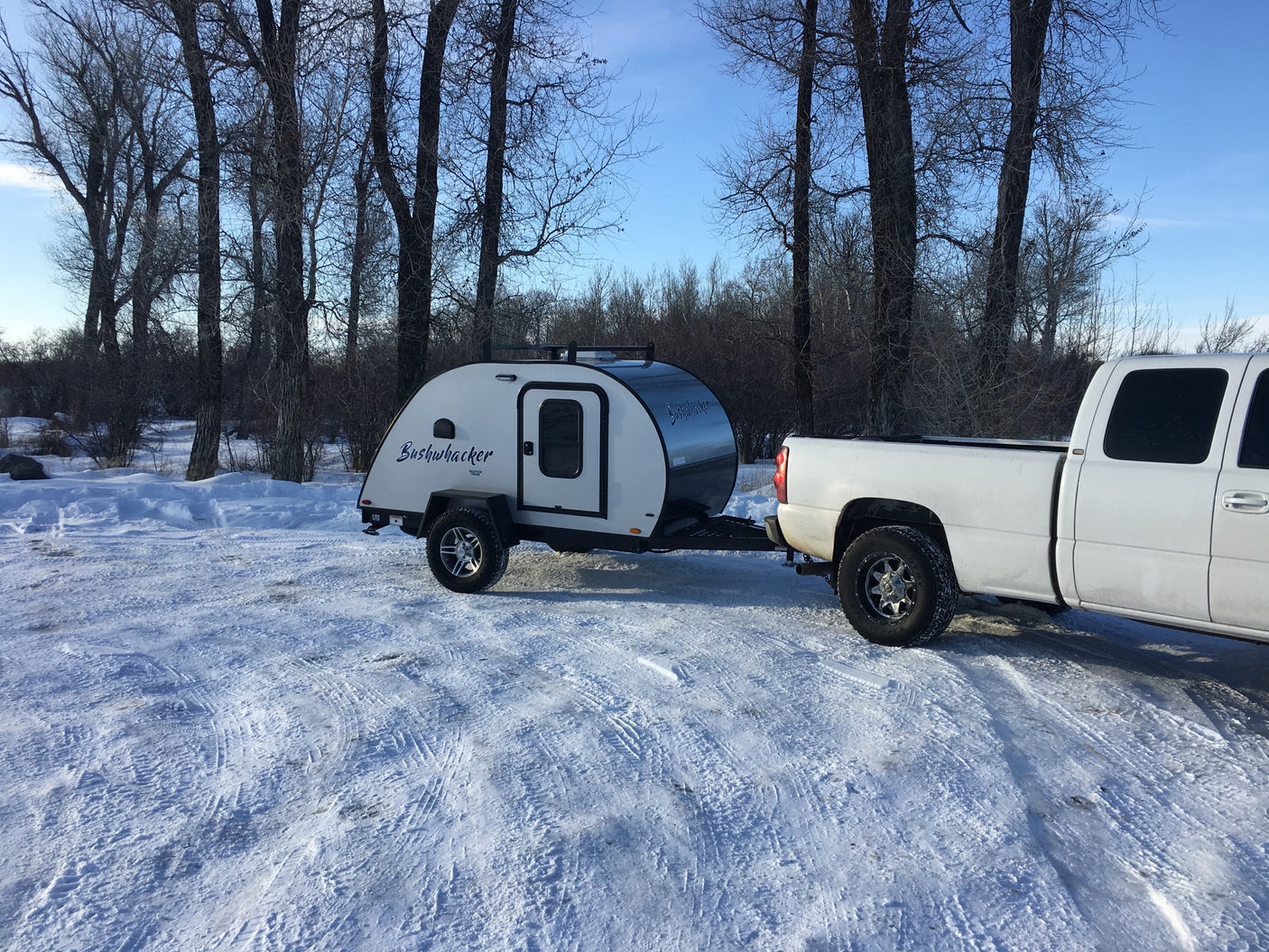 White pickup tows a Bushwhacker teardrop trailer in a snowy landscape, perfect for an rv rental adventure.