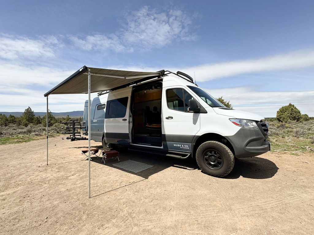 A white & gray RV rental camper van parked outdoors with an extended awning for shade.