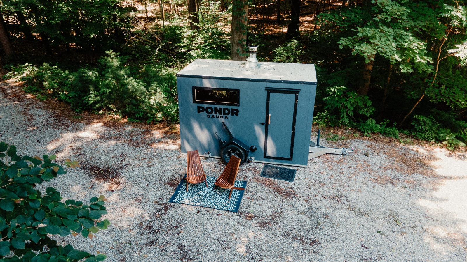 Unique rv rental: a gray mobile Pondr Sauna in a lush forest clearing. Two chairs on a patterned rug await outside.