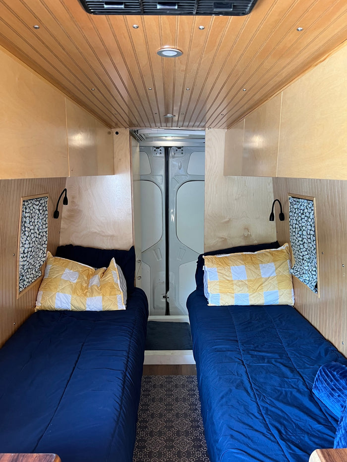 Cozy rv rental interior: two single beds with blue comforters, yellow pillows, wood paneling, and reading lamps.