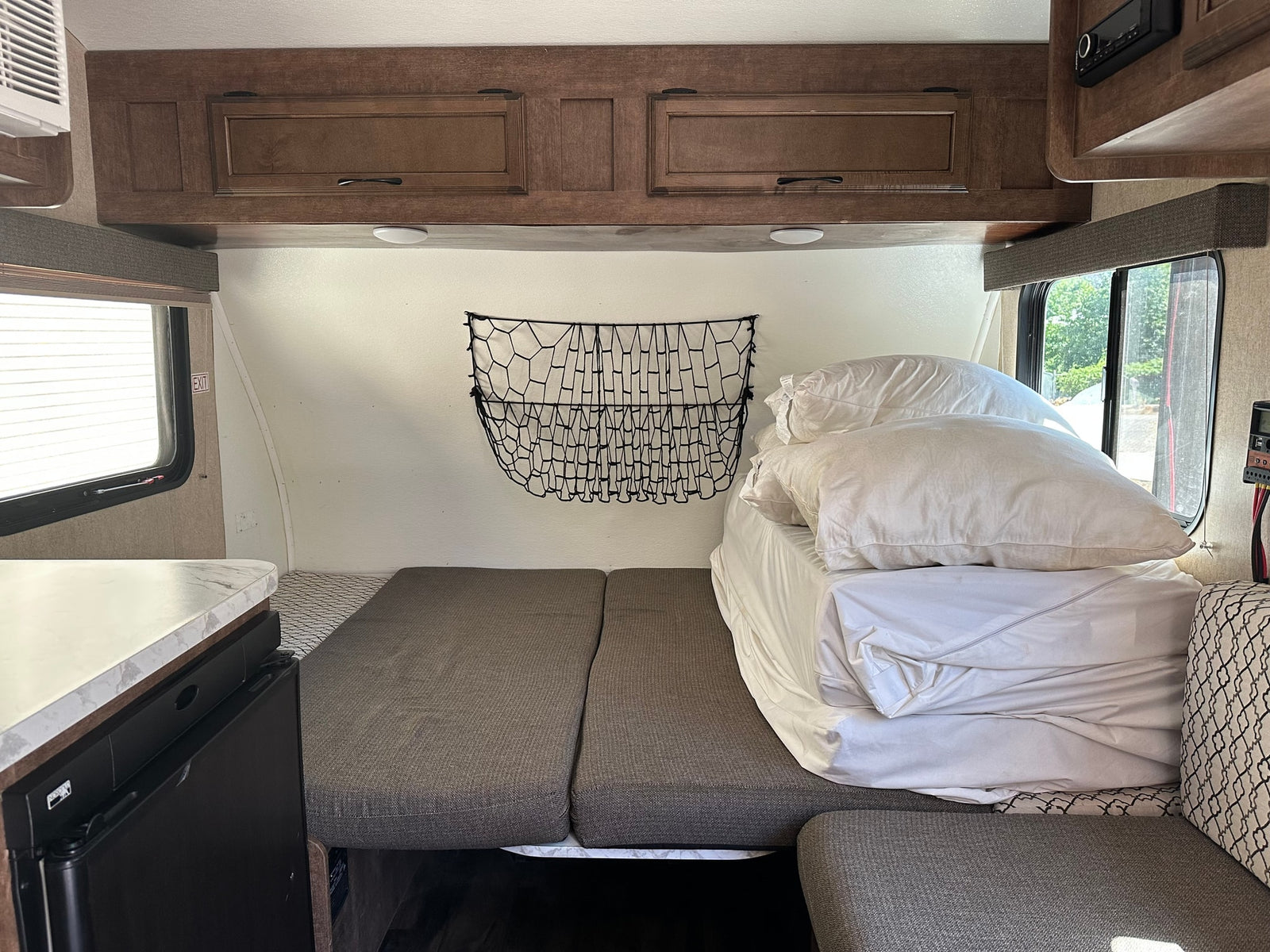 RV rental interior features a neatly made bed with white linens, wooden overhead cabinets, and a side window, ready for travel.