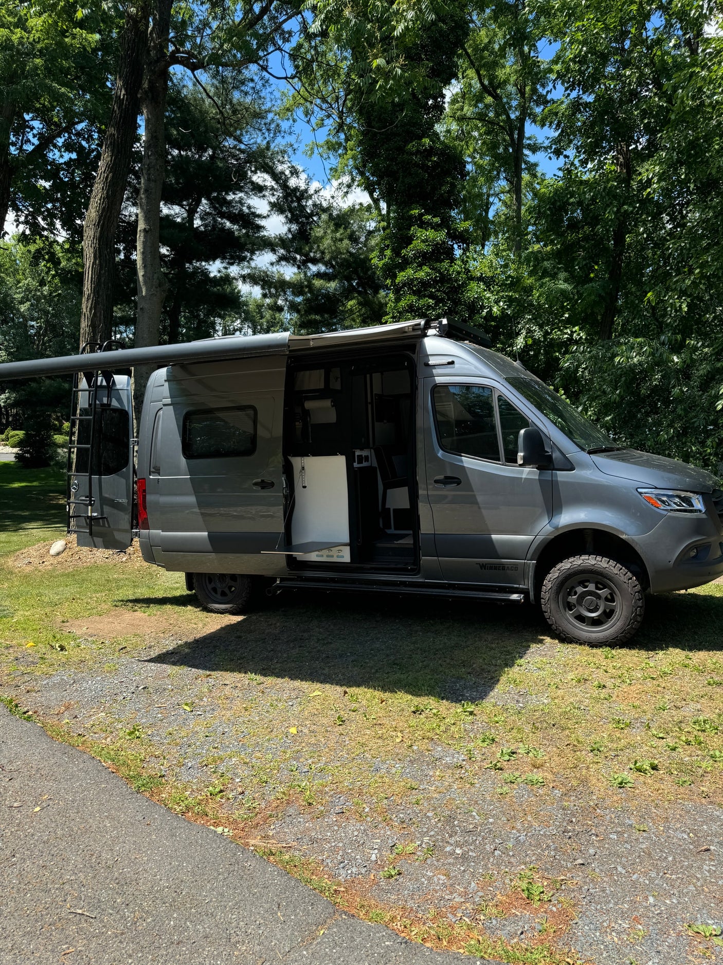 Modern gray Mercedes Sprinter RV rental with open side showing interior, parked under trees with awning extended.