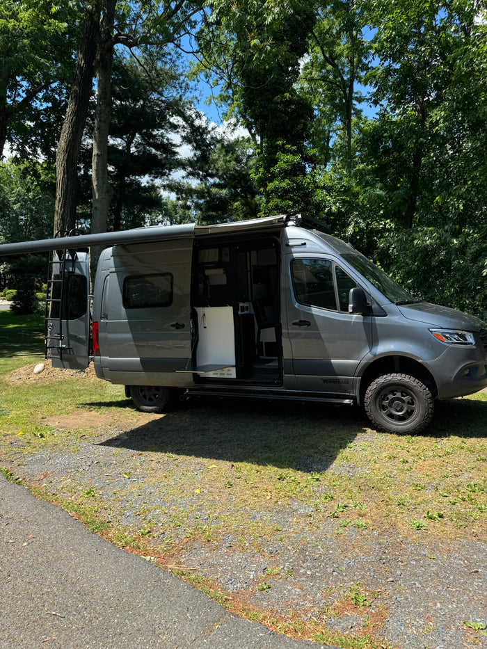 Modern gray Mercedes Sprinter RV rental with open side showing interior, parked under trees with awning extended.