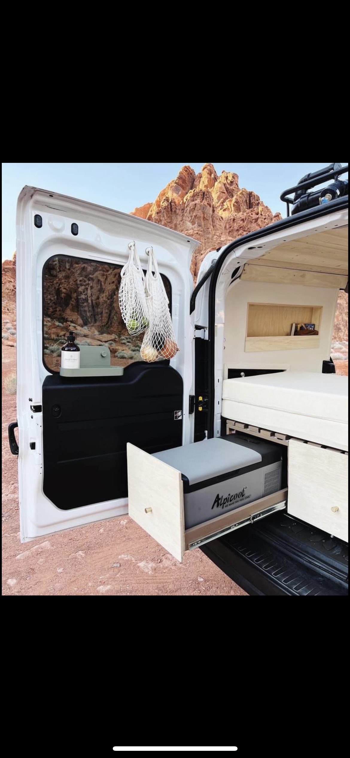 An RV rental camper van, featuring a pull-out fridge, compact kitchen setup, and bed, parked amidst red desert mountains.