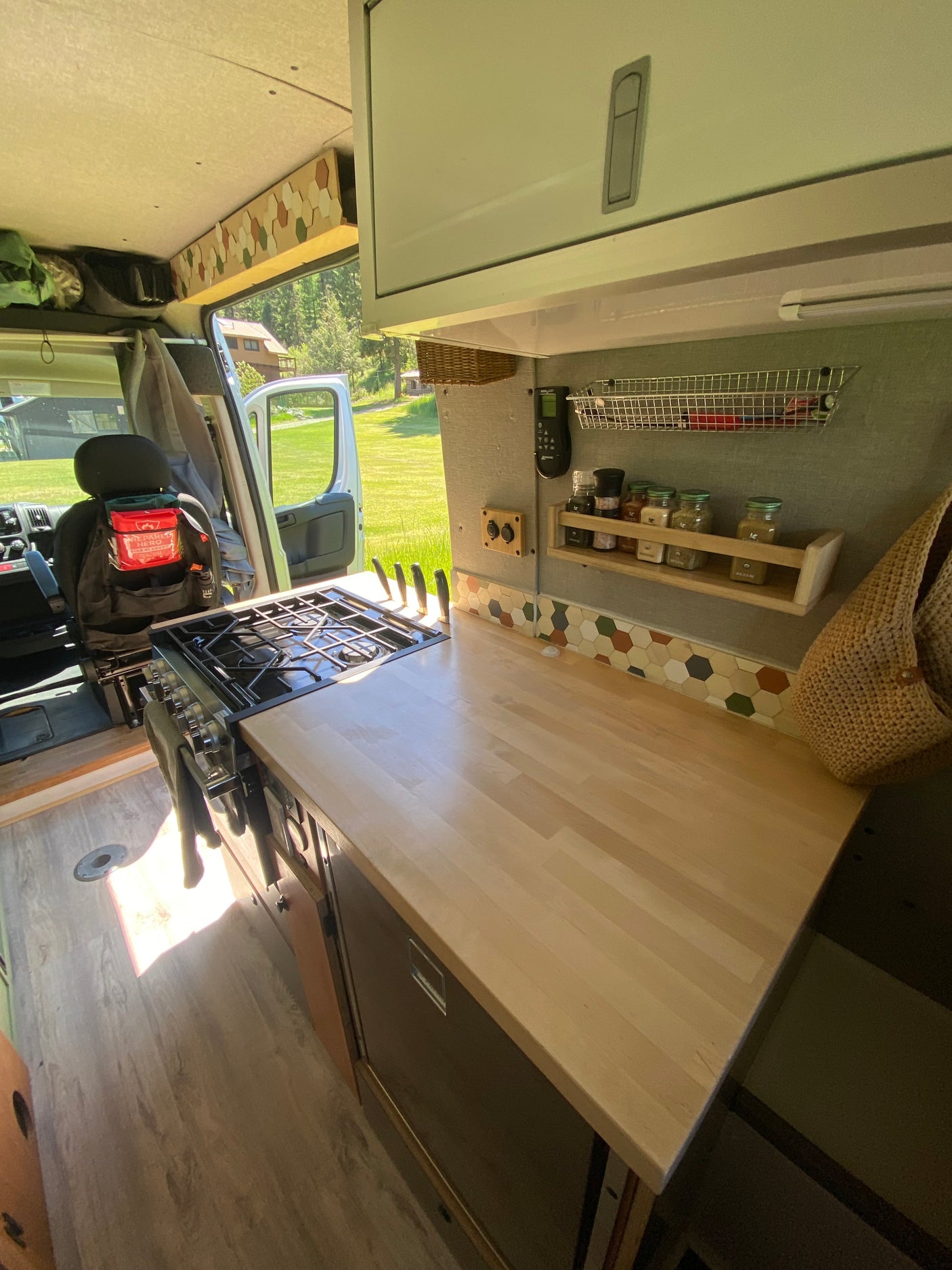 RV rental kitchen: stove, light wood counter, spice rack, & hexagonal tile backsplash. Open door shows a grassy view.