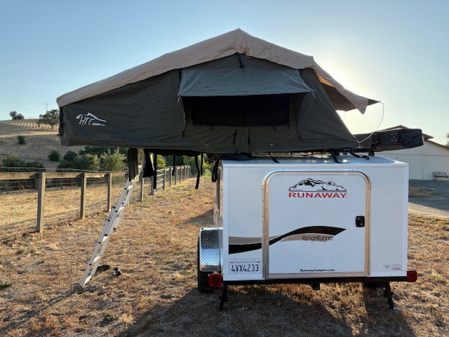 A white Runaway travel trailer with a large green rooftop tent, deployed with its ladder, ready for your rv rental adventure.
