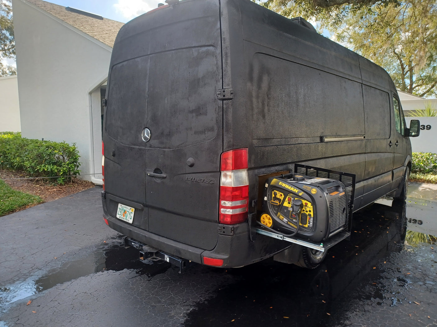 Black RV rental van with a Firman generator extended on a slide-out tray from its side compartment, parked on a wet driveway.