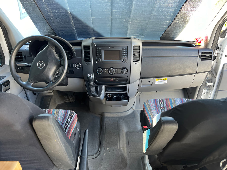 RV rental interior: View of the driver's cockpit, dashboard, and front seats with patterned covers. Sunshade on windshield.