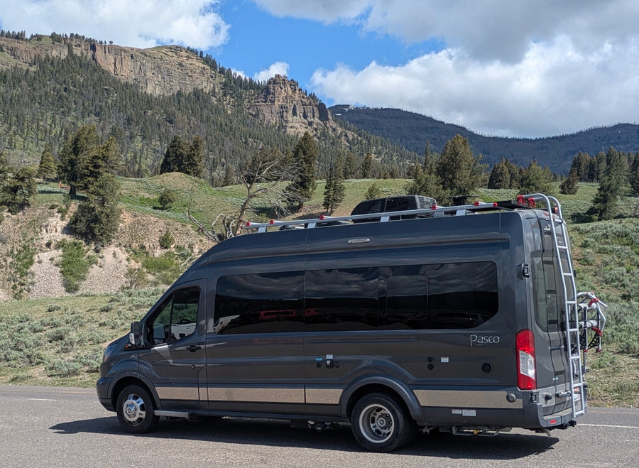 with mountains in the background under a cloudy sky.|A dark gray RV rental with a bike rack is parked on a scenic mountain road
