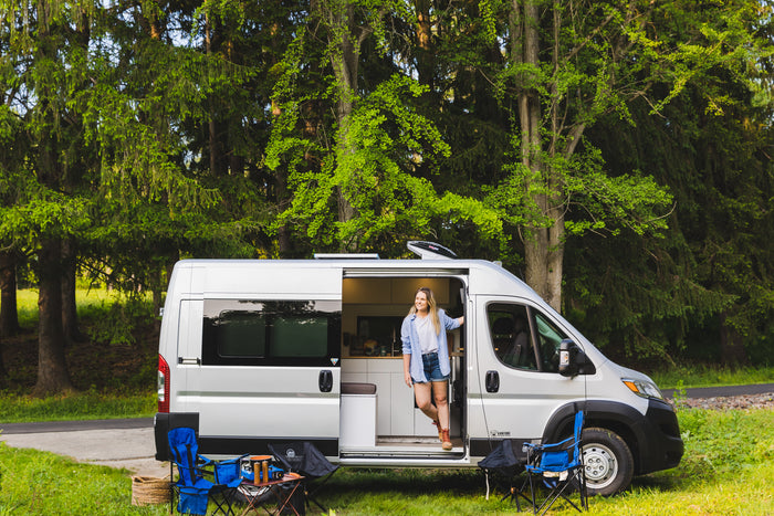 A smiling woman stands in her modern rv rental camper van, parked in a vibrant forest with camping gear ready.