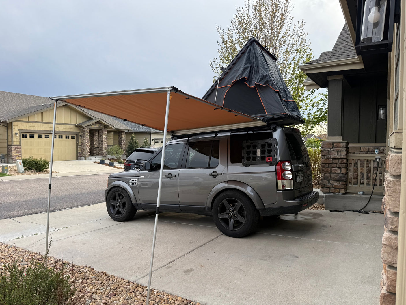 This gray SUV with a rooftop tent and awning could be an RV rental, parked in a suburban driveway.
