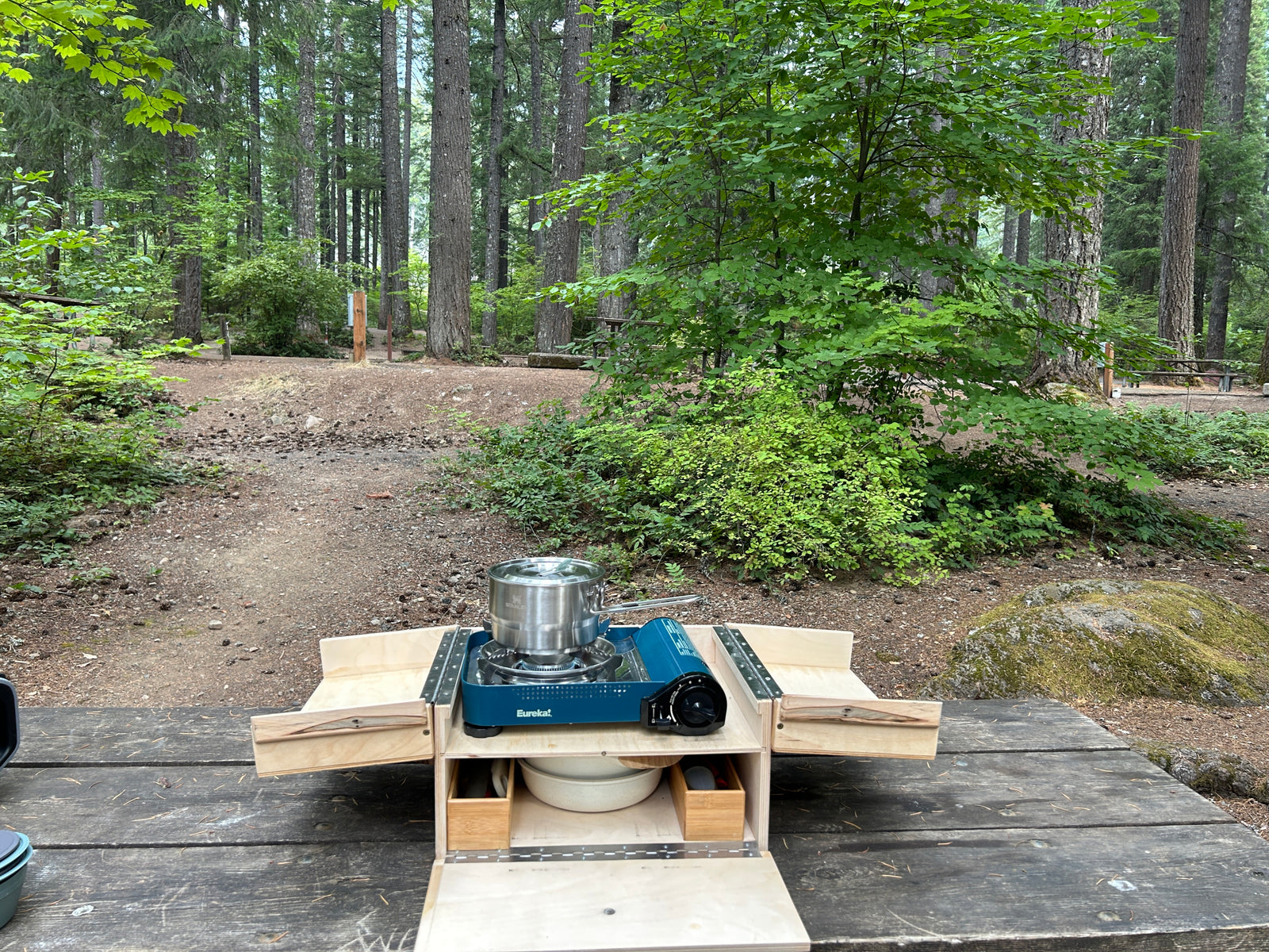 Enhance your RV rental trip! A portable camp kitchen with a stove and pot sits on a picnic table in a forest campsite.