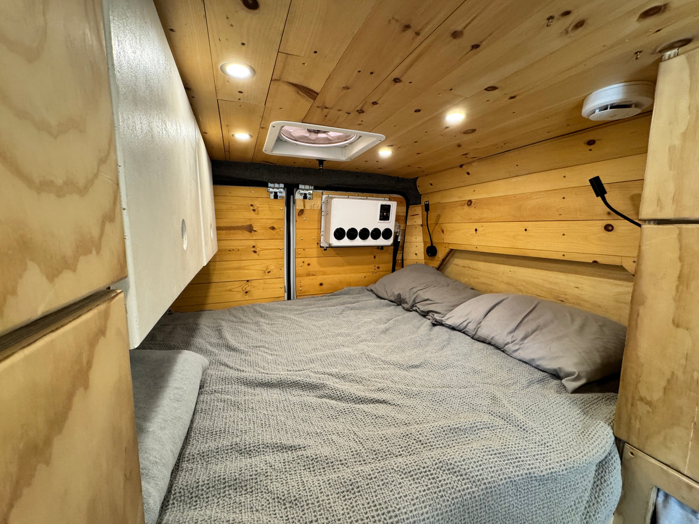 Warm RV rental bedroom with wood paneling, gray bedding, overhead lights, a roof vent, and wall-mounted controls. Left side storage.