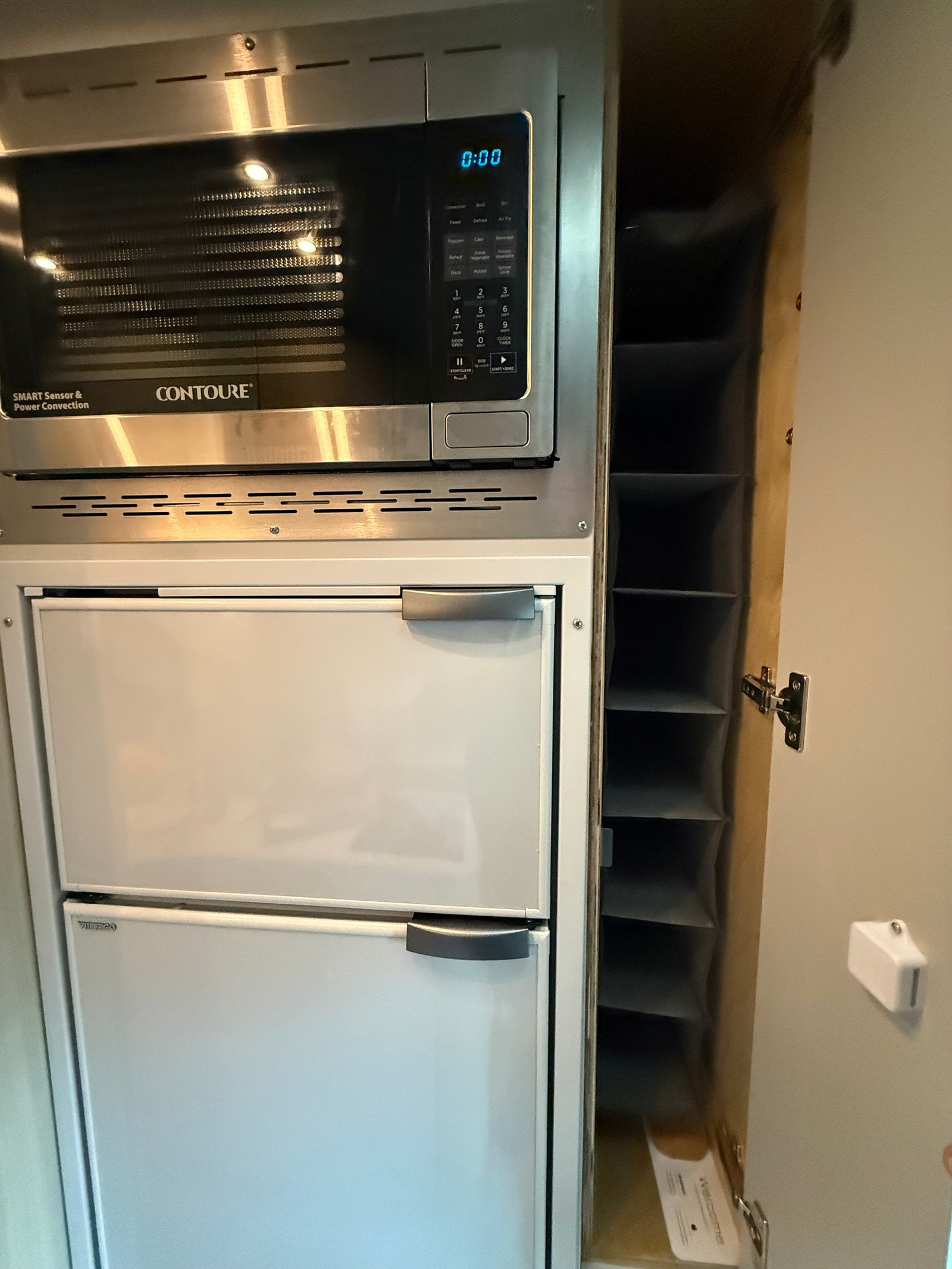 Inside an RV rental: a stainless steel microwave, two white fridge drawers below, and an open storage cabinet on the right.