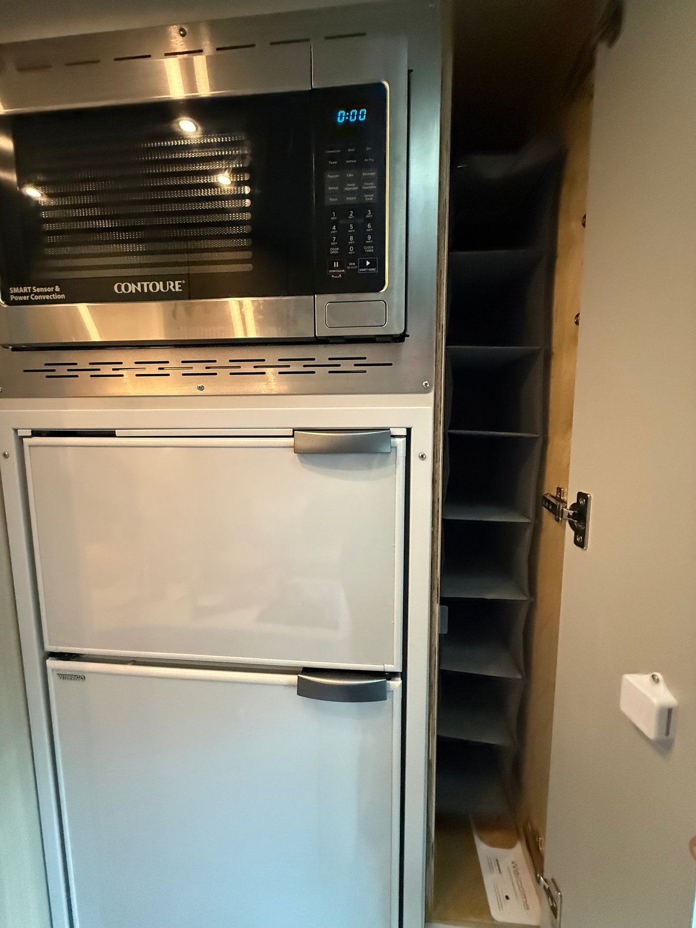 Inside an RV rental: a stainless steel microwave, two white fridge drawers below, and an open storage cabinet on the right.