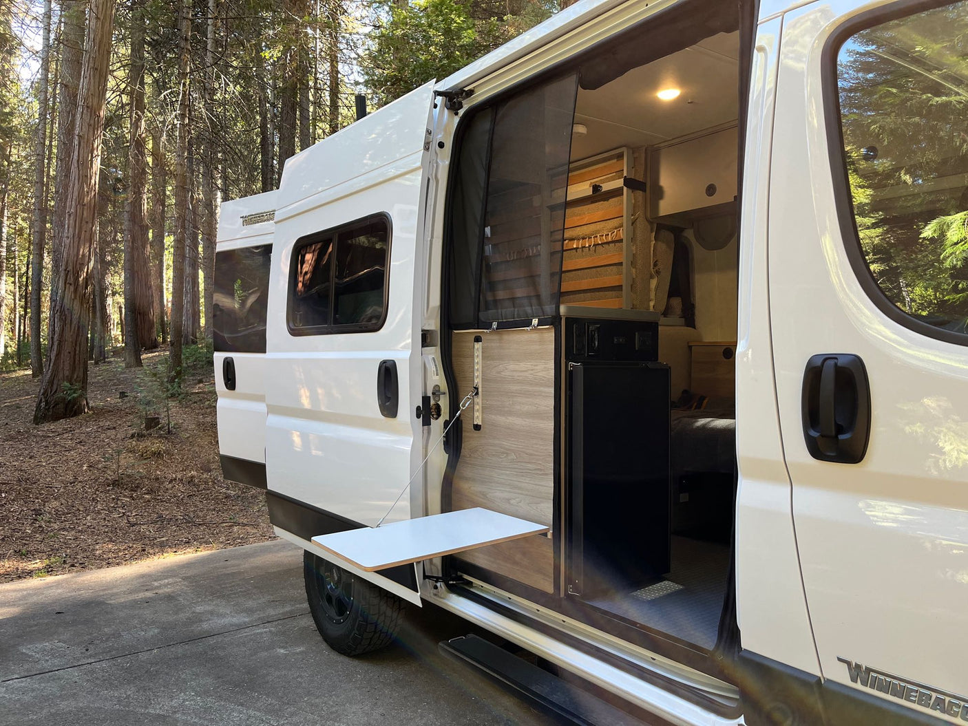Discover freedom with this RV rental. White camper van in a sunny forest, its side door open to reveal a cozy interior and pull-out table.