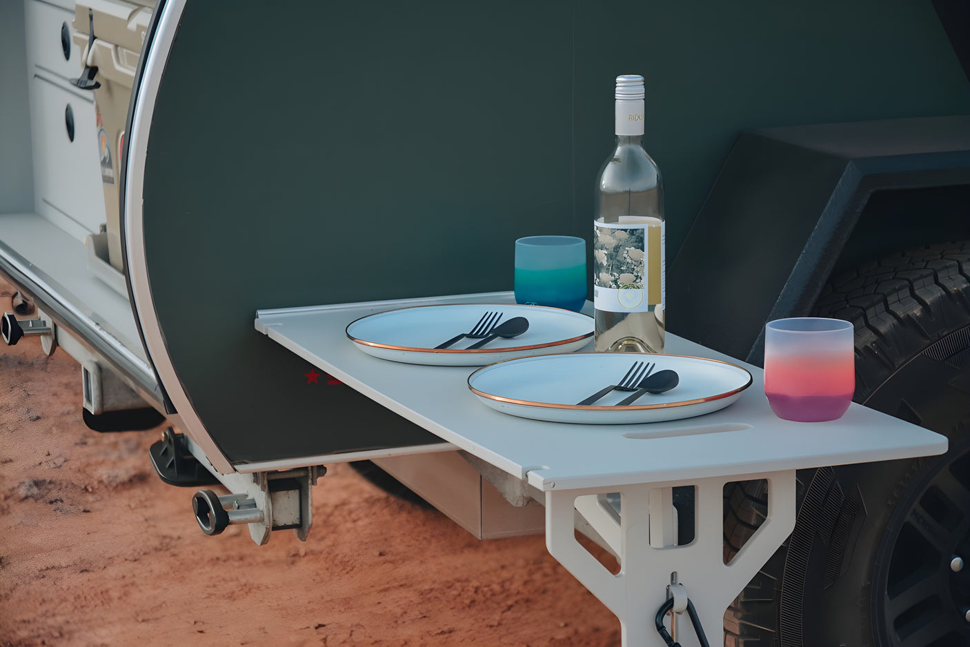 An RV rental's clever pull-out table is set for two with plates, cutlery, a wine bottle, and colorful glasses for outdoor meals.